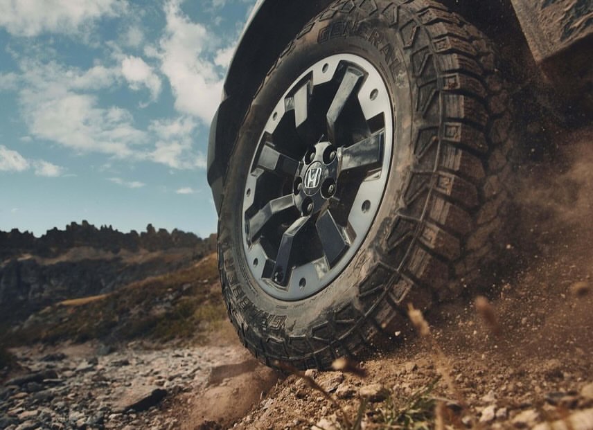 Close-up of a Honda vehicle's tire on a rocky, dirt terrain under a sky with scattered clouds, emphasizing off-road capabilities.