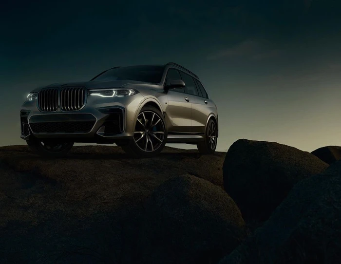 Silver BMW luxury SUV parked on rocky terrain under a dark twilight sky with illuminated headlights.