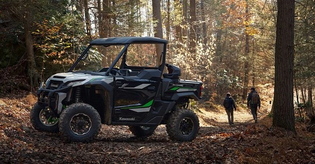 A black and green Kawasaki side-by-side ATV is parked on a leafy forest path with two people walking in the distance during daytime.