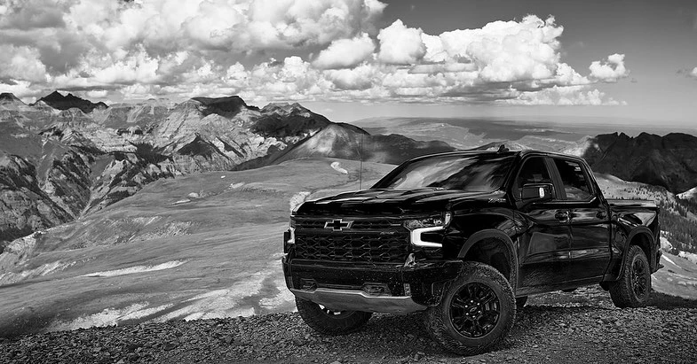 Black Chevrolet pickup truck parked on rocky terrain, with a mountainous landscape and cloudy sky in the background, monochrome shot.