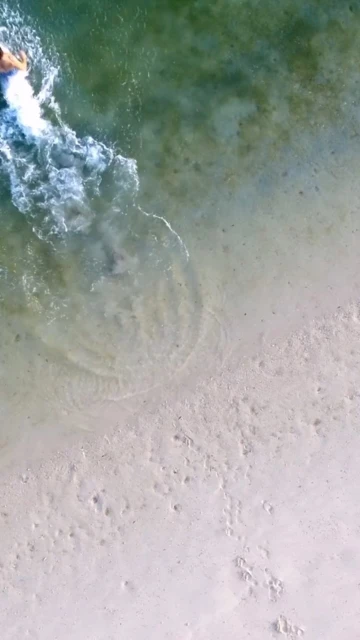 Aerial shot of an adult in clear ocean water near a sandy beach, with visible ripples and the sandy seabed.