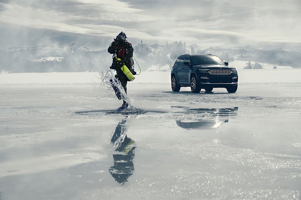 A diver emerges from an icy lake alongside a parked black SUV on a winter day with snow-covered mountains in the background.