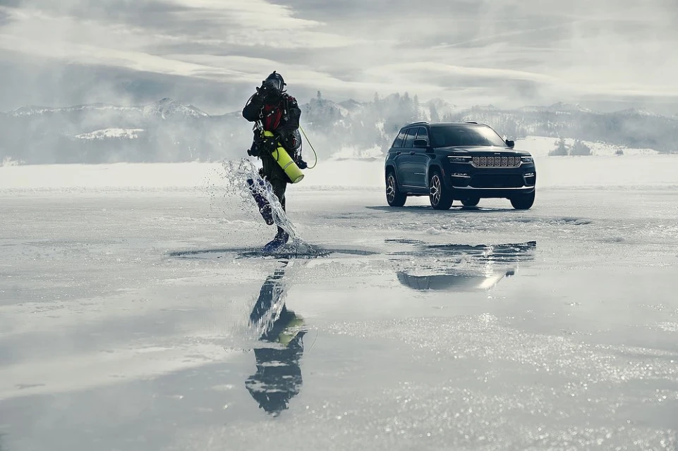 A diver emerges from an icy lake alongside a parked black SUV on a winter day with snow-covered mountains in the background.