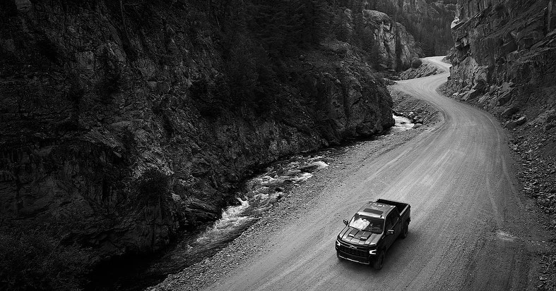 Black pickup truck driving on a dirt road between rocky cliffs and a river in a remote, monochrome setting.
