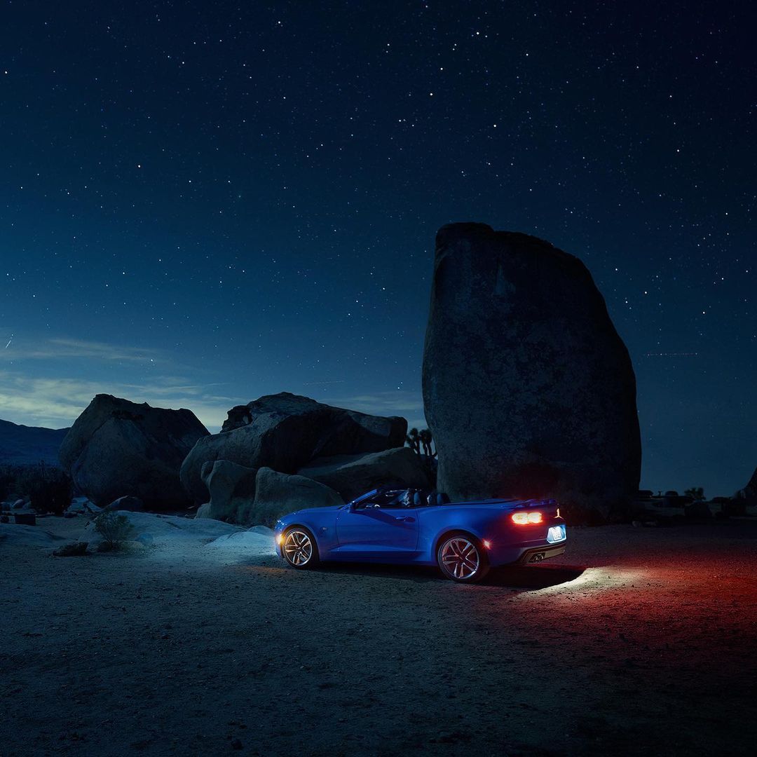Blue convertible car parked in a desert landscape at night, with large rock formations and a starry sky in the background.