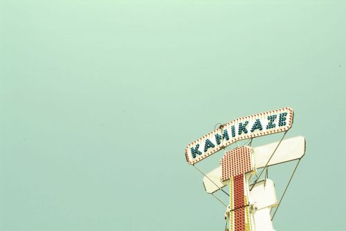 Kamikaze ride sign against a pale blue sky, featuring a white structure with red accents and string supports.