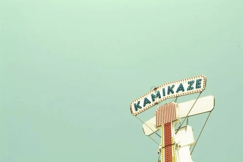 Kamikaze ride sign against a pale blue sky, featuring a white structure with red accents and string supports.