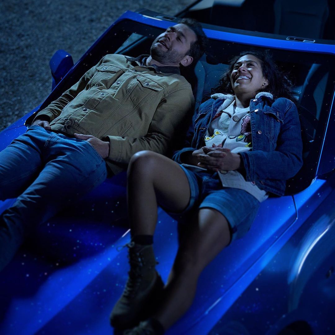 A smiling man and woman lying on the hood of a blue car at night. The couple is relaxing and enjoying the peaceful moment outdoors.