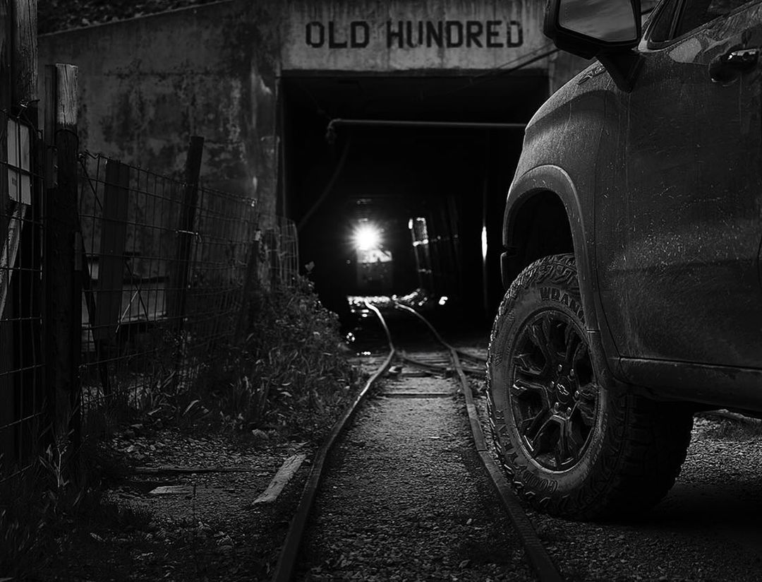 Grayscale image of a truck parked near the entrance of the Old Hundred Gold Mine, showcasing its tire and rugged exterior in a dimly lit setting.