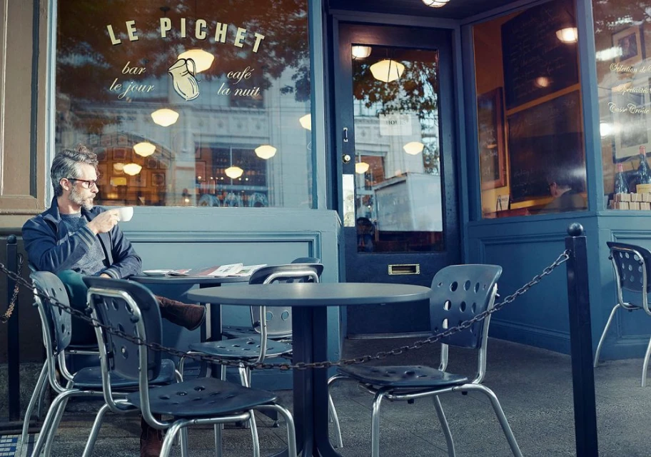Man with glasses enjoys coffee at outdoor cafe table near French restaurant Le Pichet in a city setting with chairs and newspapers.