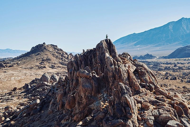 Lone hiker stands atop a rocky peak in the Alabama Hills, with distant mountains under a clear sky, showcasing the stark desert landscape.