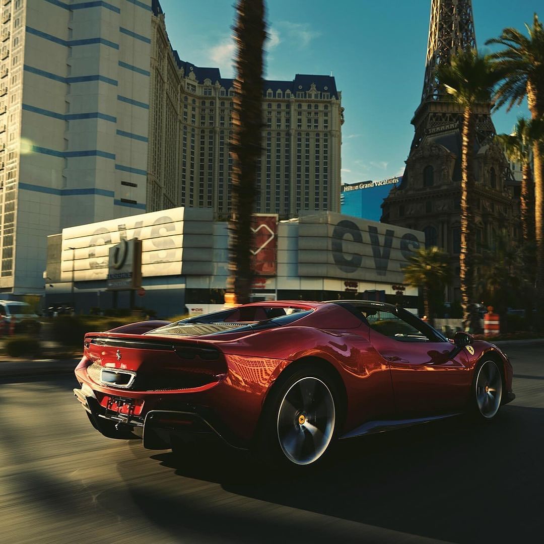 Red Ferrari sports car drives down Las Vegas strip on a sunny day, with the Eiffel Tower replica and palm trees visible in the background.