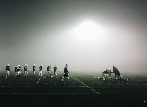 American football team practices at night in heavy fog under bright stadium lights; multiple players on a marked field wearing helmets.