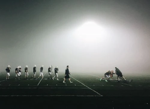 American football team practices at night in heavy fog under bright stadium lights; multiple players on a marked field wearing helmets.