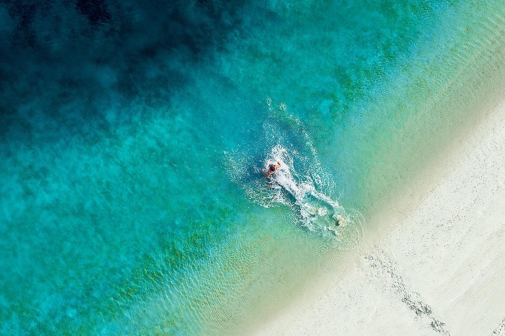 Aerial shot of a person swimming in clear turquoise ocean water next to a white sand beach on a bright sunny day, creating splash.