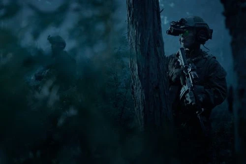 Two military soldiers in a dark forest at night wearing full gear including night vision goggles and holding rifles, standing guard.