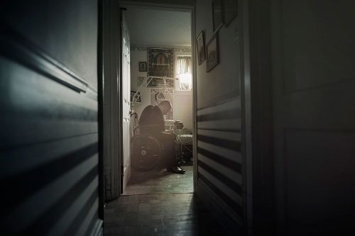 Low-key shot of elderly person in wheelchair in dimly lit room with doorway. Interior setting with artwork and architectural elements.