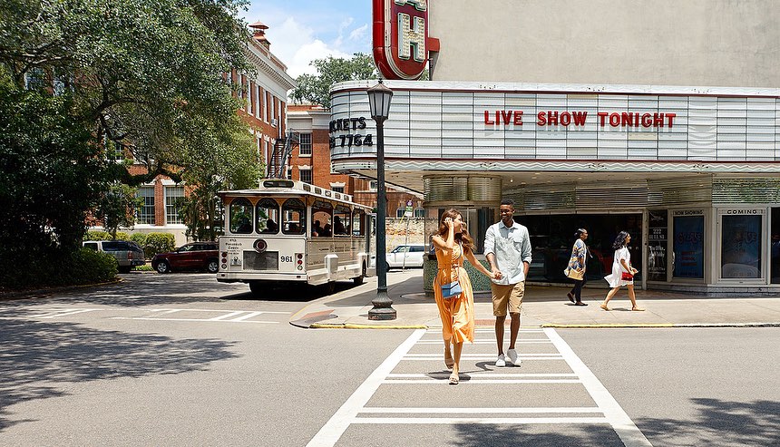 A couple holds hands while crossing a street in front of a vintage trolley and theater with trees and brick buildings in the background.