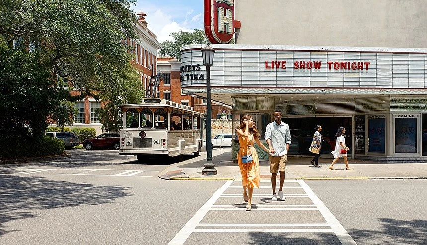 A couple holds hands while crossing a street in front of a vintage trolley and theater with trees and brick buildings in the background.