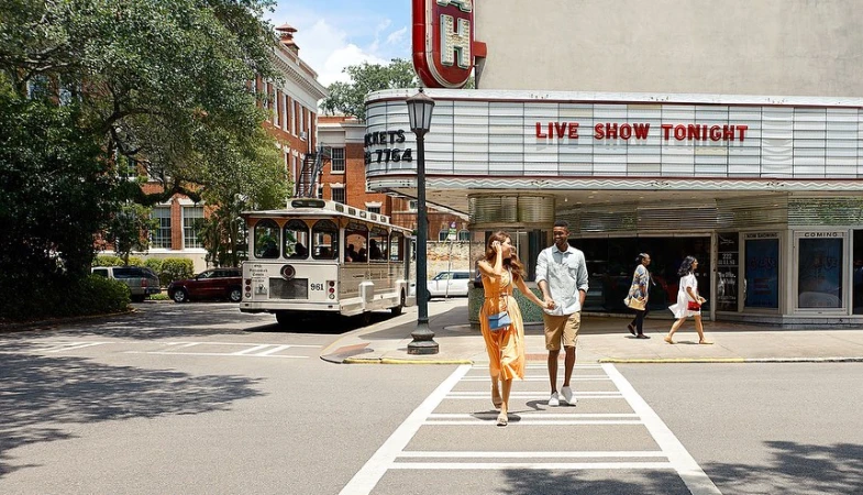 A couple holds hands while crossing a street in front of a vintage trolley and theater with trees and brick buildings in the background.