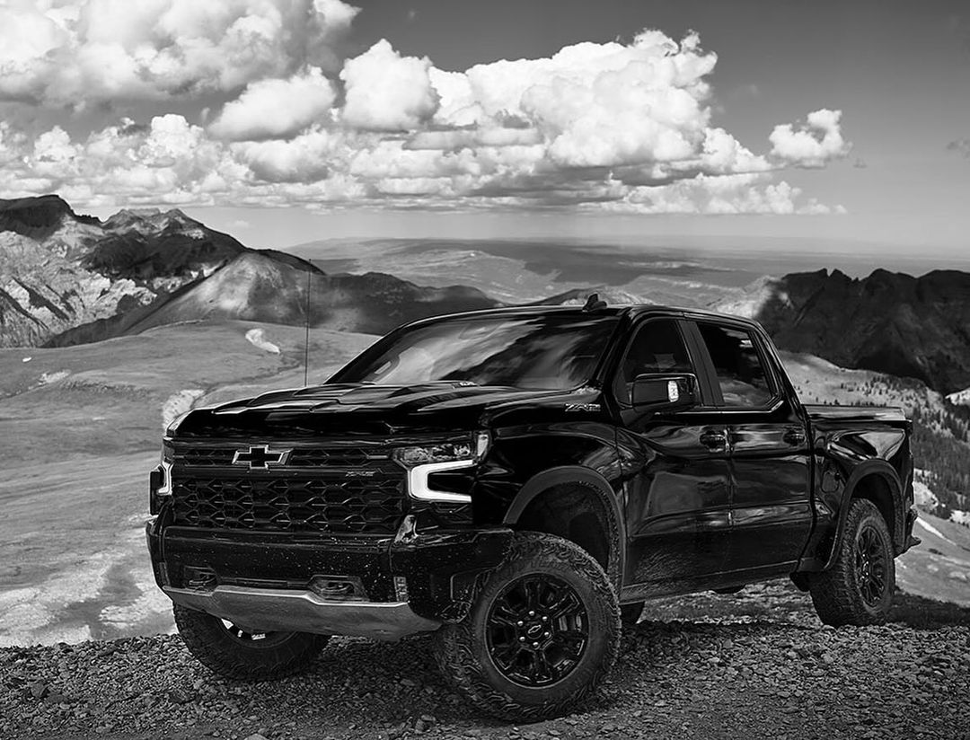 Black Chevrolet pickup truck parked on a hilltop with distant mountains and cloudy skies providing a scenic backdrop in black and white.