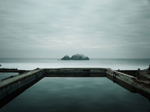 Ocean water fills concrete pool on overcast day with distant island. Long exposure creates smooth water surface with reflective surface.