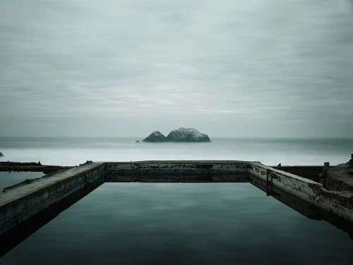 Ocean water fills concrete pool on overcast day with distant island. Long exposure creates smooth water surface with reflective surface.