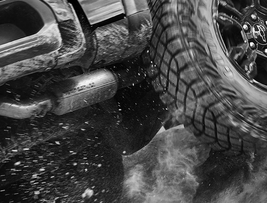 Black and white shot of a truck tire in motion with water droplets visible. The exhaust pipe is visible with water spraying.