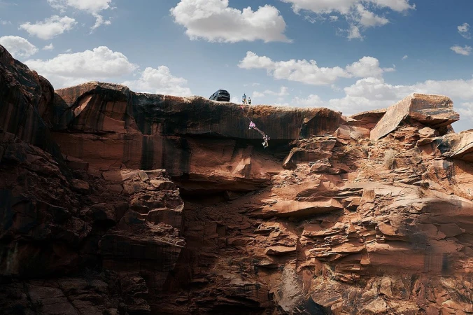 BASE jumper leaps from the edge of a red rock cliff on a rope, a car and several people stand in the background against a partly cloudy sky.
