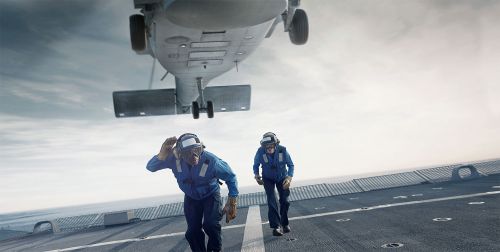 Two aviation personnel run on the deck of an aircraft carrier as a helicopter approaches for landing with cloudy sky background.