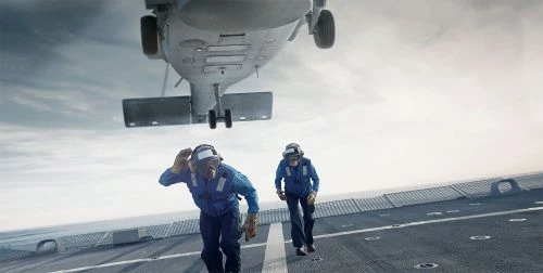 Two aviation personnel run on the deck of an aircraft carrier as a helicopter approaches for landing with cloudy sky background.