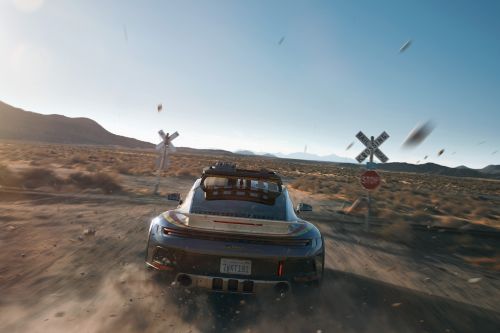 An all-terrain sports car drives along a dusty desert road with a railroad crossing sign in the background under a clear blue sky.
