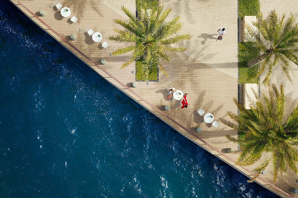 Aerial view of people relaxing on a waterfront promenade with palm trees and white patio furniture beside calm blue water on a sunny day.