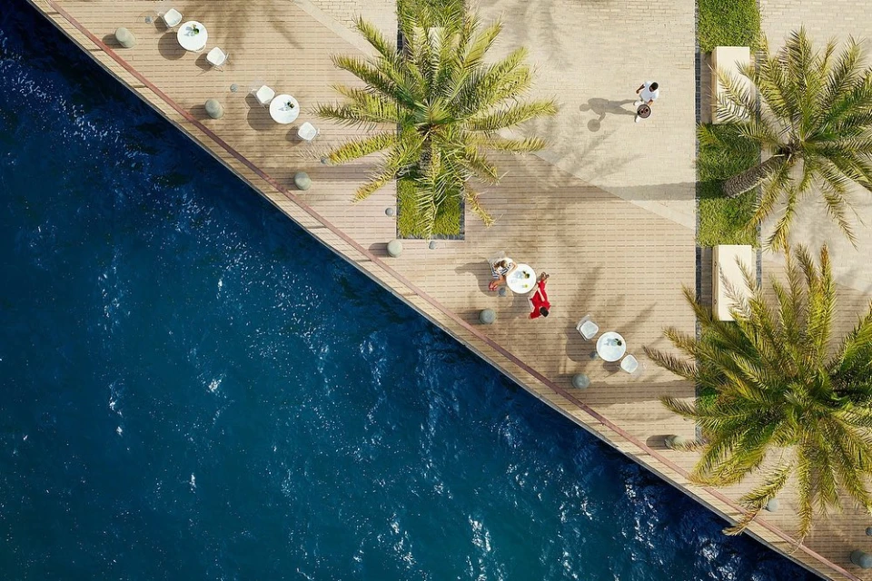 Aerial view of people relaxing on a waterfront promenade with palm trees and white patio furniture beside calm blue water on a sunny day.