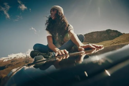 A woman wearing a beanie and dark vest sitting cross-legged on the roof of a car under a bright sky, photographed by Dana Neibert.