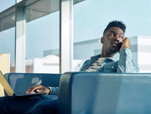 Young adult man sits in blue airport lounge chair using laptop. He wears a blue corduroy shirt and looks away from screen in thought.