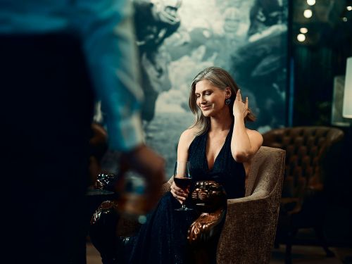 Elegant mature woman in a black dress sits in a chair, holding a wine glass, with a blurred figure in the foreground in a dark indoor setting.