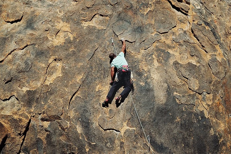 Adult male rock climbing up a textured rock face wearing a teal t-shirt, black pants, and climbing shoes; outdoor sport in natural sunlight.