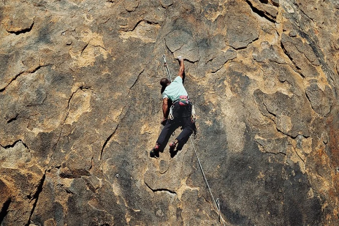 Adult male rock climbing up a textured rock face wearing a teal t-shirt, black pants, and climbing shoes; outdoor sport in natural sunlight.