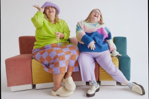 Two plus-size women pose in brightly colored knitwear on a vibrant couch in a studio, laughing and gesturing with their hands.