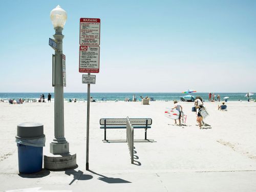 Beach scene with a lamppost, warning signs, a trash can, and distant figures on sandy shore under clear blue sky.