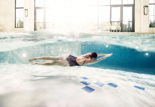 Underwater shot of an adult female swimmer in a dark swimsuit, goggles, and swim cap doing the butterfly stroke in an indoor pool.