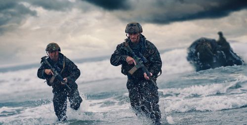 Two armed soldiers in camouflage run through ocean surf, facing forward, against a cloudy sky and rock formations.