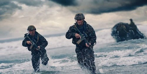 Two armed soldiers in camouflage run through ocean surf, facing forward, against a cloudy sky and rock formations.