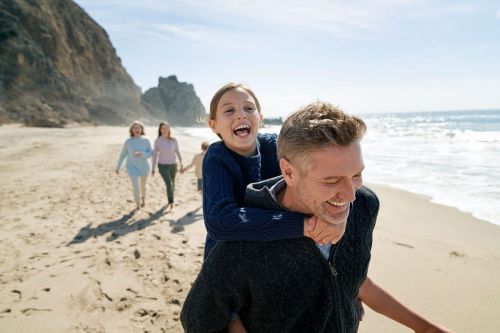 Father gives daughter a piggyback ride on a sandy beach under a sunny sky, with other family members walking in the background.