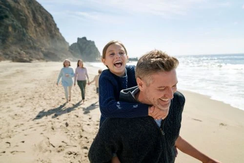 Father gives daughter a piggyback ride on a sandy beach under a sunny sky, with other family members walking in the background.