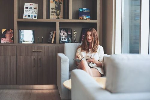 Woman with long hair in light jacket sits in an armchair reading a book, in a living room with bookshelves and a large window.