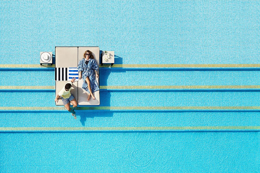 Aerial view of a couple relaxing on a daybed next to a swimming pool, featuring clean architectural lines and bright, natural lighting.