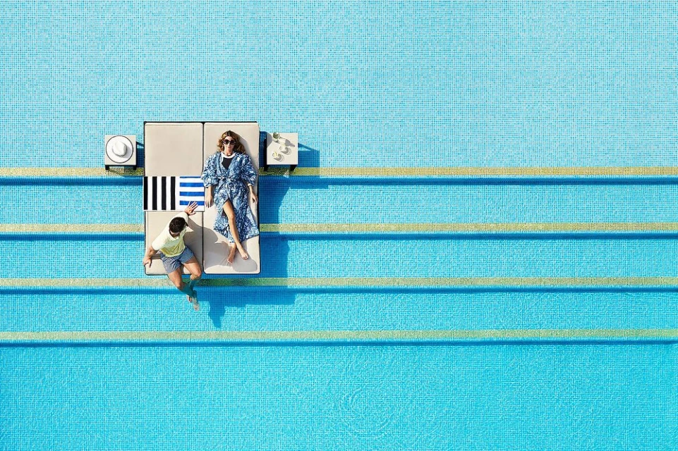 Aerial view of a couple relaxing on a daybed next to a swimming pool, featuring clean architectural lines and bright, natural lighting.