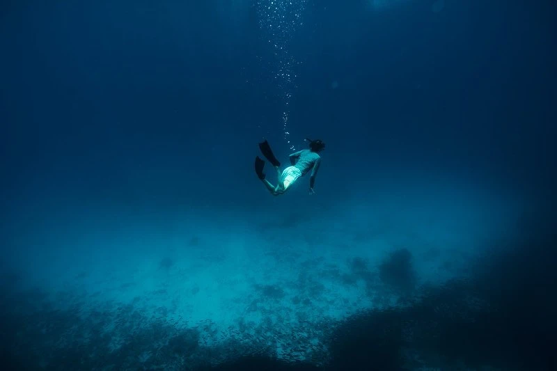 Underwater view of a person swimming in dark blue water with bubbles rising above, with a coral reef visible below.